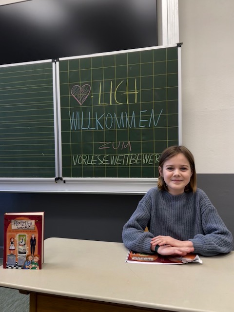 Ein Mädchen sitzt vor einer Tafel, Aufschrift: Herzlich Willkommen zum Vorlesewettbewerb